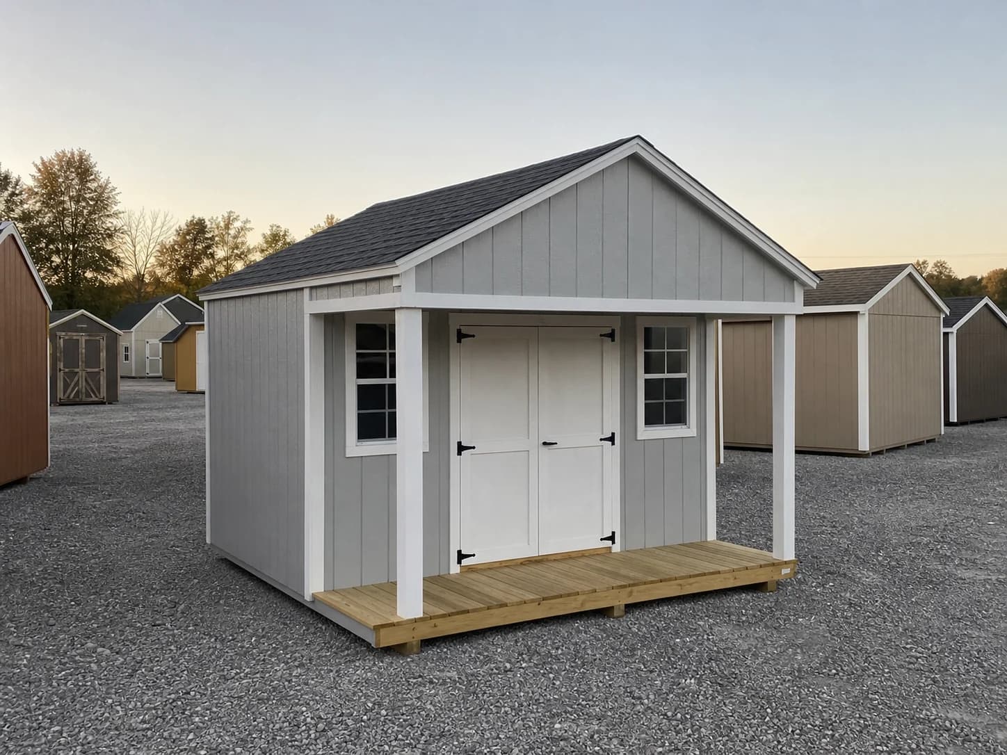 A 12 by 16 cottage shed with a porch in the front row of a gravel display lot, with the ten or twelve buildings behind it that stand in for a range making thousands.